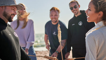 Eine Menschengruppe sthet um eine Feuerstelle und grillen Stockbrot.