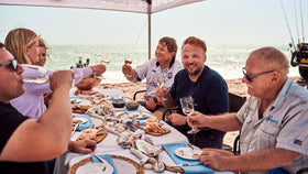 Eine Gruppe Menschen sitz in freier Natur um einen gedeckten Tisch und genießen das Essen.