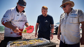 Drei Männer stehen am Strnd um einen Grill. Einer belegt den Rost mit eir Falafel ähnlichen Paste.