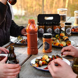 Gewürzstreuer mit Magic Dust Rub auf einem Holztisch neben Tellern mit Grillgut.